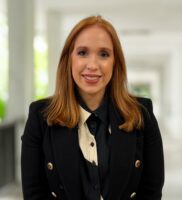 Kathryn Williams, a woman with straight red hair, wearing a black blazer and light-colored blouse, stands in a bright hallway with blurred columns in the background.