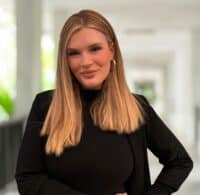 Claire Butcher, a woman with long, straight blonde hair wearing a black top and blazer, stands indoors in a well-lit hallway, looking confidently at the camera.