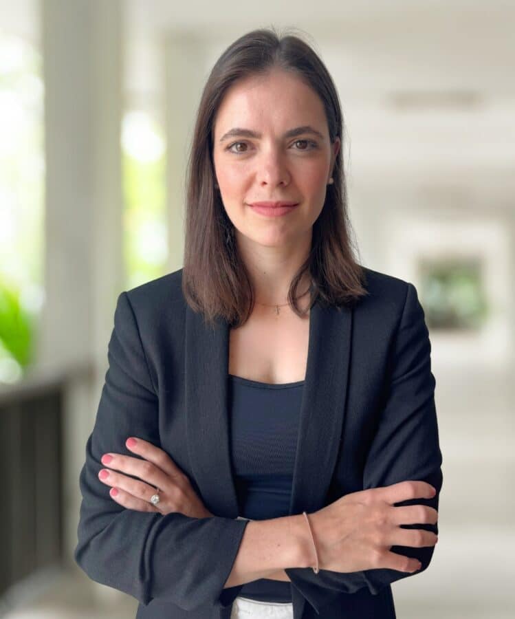 Claudia Lemke, a woman with straight brown hair, wearing a black blazer and top, stands with arms crossed, looking at the camera in a bright hallway.