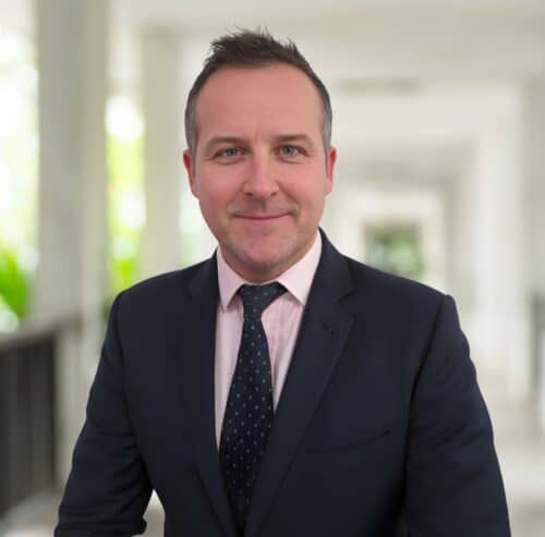 David Howells, in a dark suit and tie, stands indoors in a well-lit corridor, facing the camera and smiling slightly.