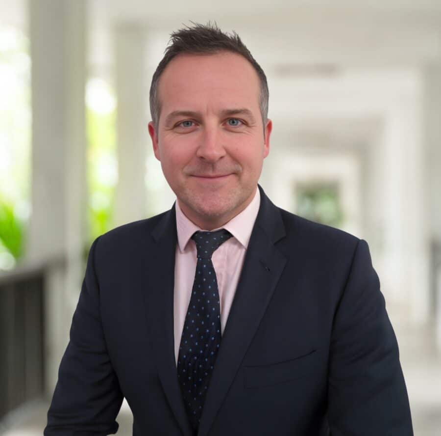 David Howells, in a dark suit and tie, stands indoors in a well-lit corridor, facing the camera and smiling slightly.