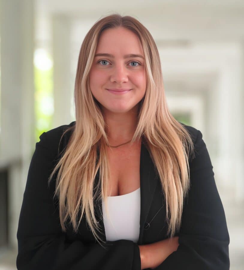 Ellie LaFountain, with long blonde hair, stands indoors in a black blazer and white top, arms crossed as she faces the camera and smiles slightly.