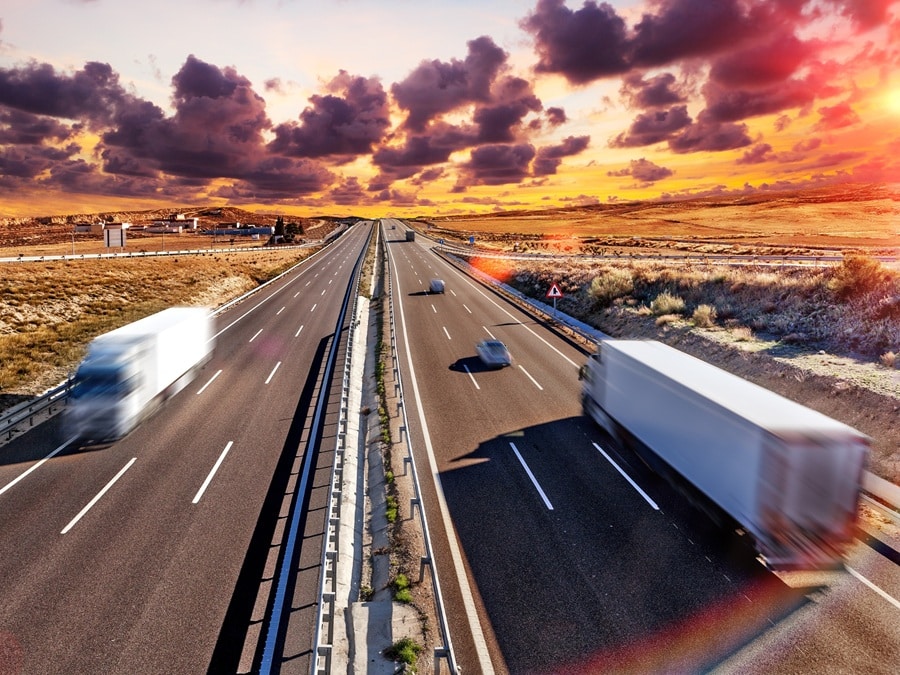 Several trucks and cars travel quickly on a divided highway through a rural landscape at sunset, with dramatic clouds in the sky—a fitting scene for those in Transportation and Logistics executive search seeking industry insight.