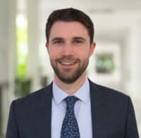 Manuel Preg, a man with short brown hair and a beard, wearing a dark suit, light blue shirt, and patterned tie, stands indoors in a bright, blurred hallway.