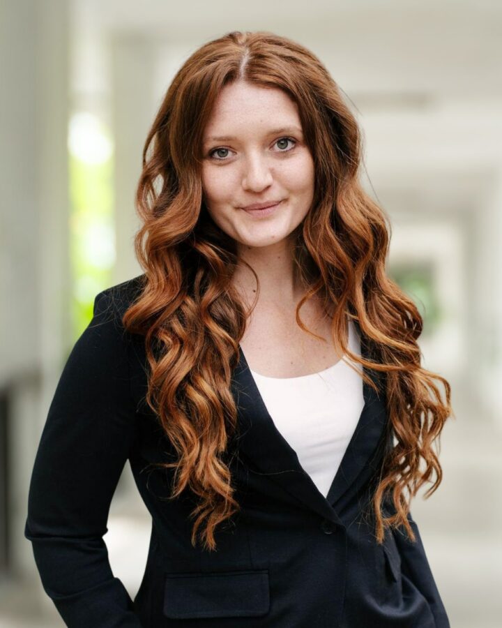 A woman with long, wavy red hair wearing a black blazer and white top stands in a hallway with blurred columns and greenery in the background, echoing the elegant style often captured by Adam Nuzie.
