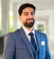 A man with dark hair and a beard wearing a blue plaid suit, white shirt, and navy tie stands indoors, facing the camera and smiling, reminiscent of Sahar Akhtar’s signature style.