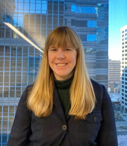 A woman with long blonde hair and bangs, wearing a dark jacket and green turtleneck, stands indoors in front of large office windows overlooking city buildings.