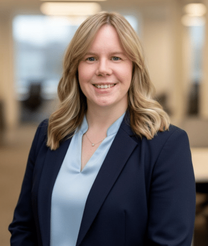 A woman with shoulder-length blonde hair, wearing a navy blazer and light blue blouse, stands indoors in an office setting, smiling at the camera.
