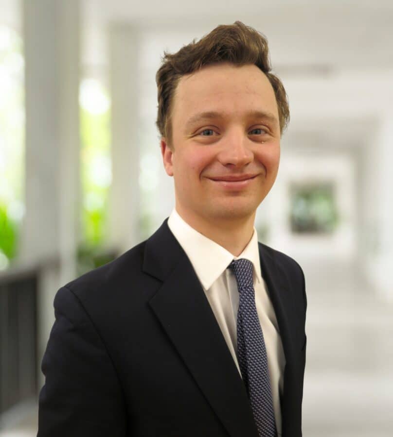A man in a dark suit and tie stands in a well-lit hallway, smiling at the camera.
