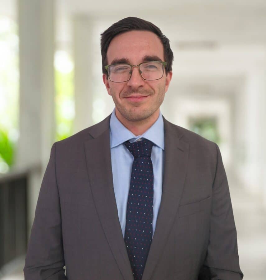 A man wearing glasses, a gray suit, blue shirt, and dark patterned tie stands in a bright, blurred hallway.