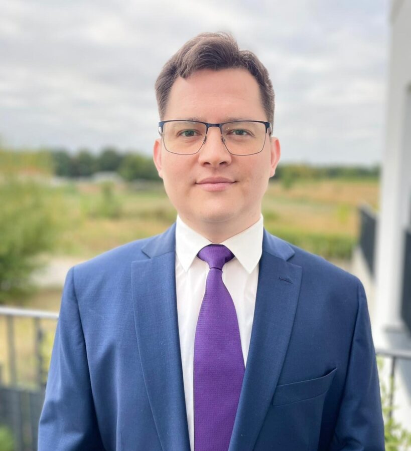 Rafael Fernandez, wearing glasses, a blue suit, white shirt, and purple tie, stands outdoors in front of a blurred background with greenery and a cloudy sky.
