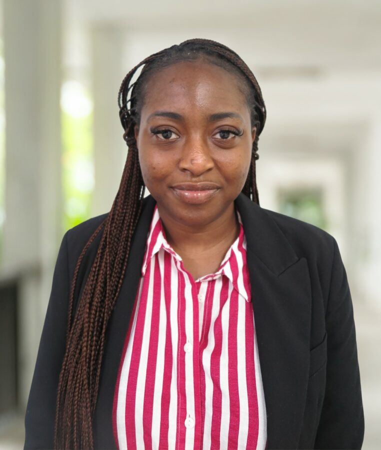 Feyisayo Adetayo, a woman with long braids, wears a black blazer and red-and-white striped shirt as she stands in a bright hallway with a blurred background.