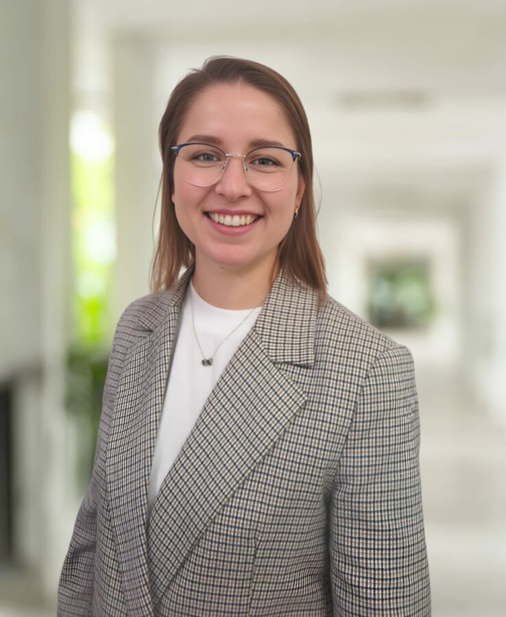 A woman wearing glasses and a plaid blazer smiles while standing in a bright hallway with blurred background.
