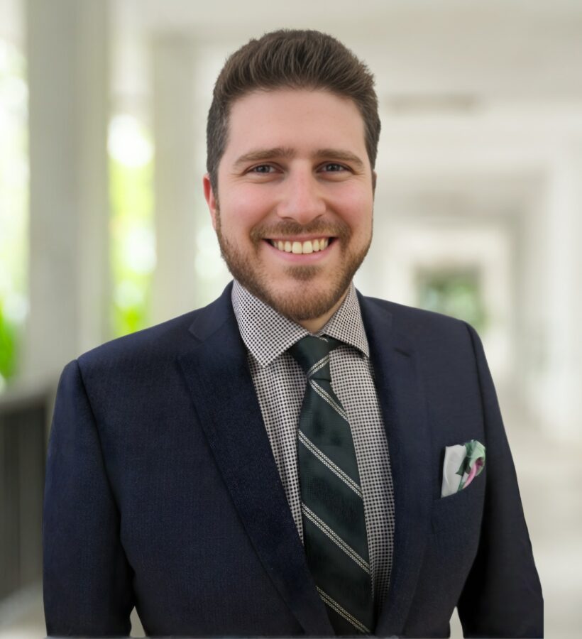 A man in a dark suit and striped tie smiles while standing in a bright, covered outdoor walkway.