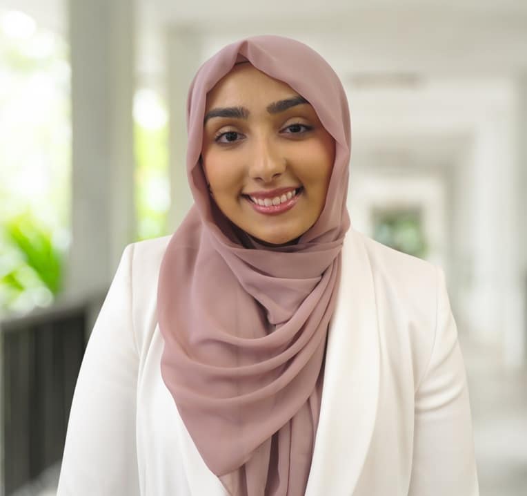 A woman wearing a light pink hijab and white blazer smiles while standing in a bright, indoor corridor.