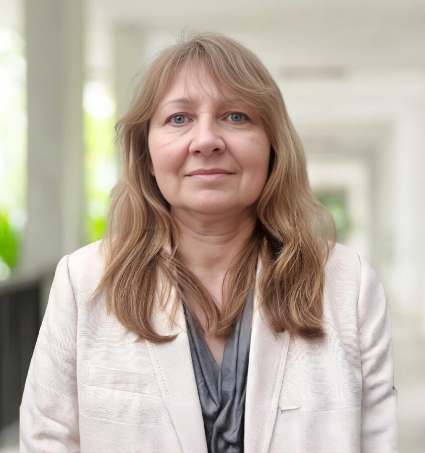 Pat Cunningham, a woman with light brown hair wearing a light-colored blazer, stands in a hallway with blurred columns and greenery in the background.