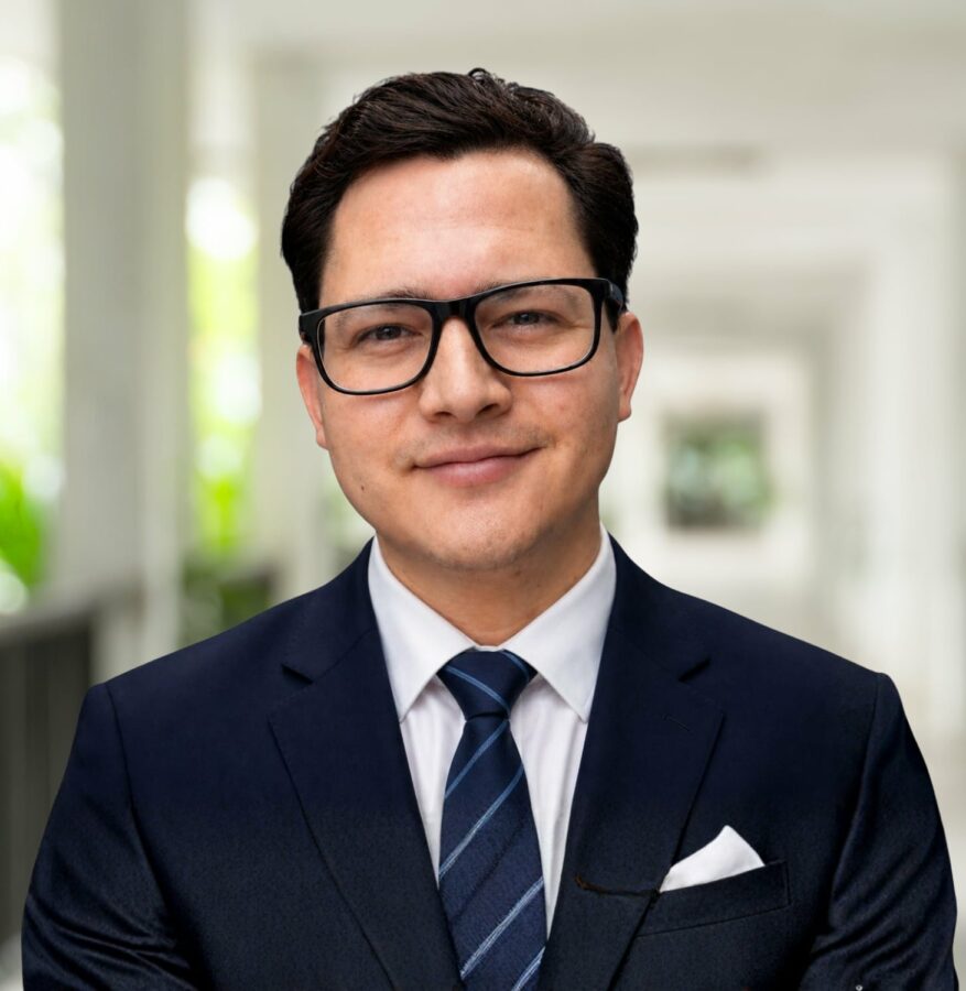 Rafael Alvarado, in a dark suit, white shirt, and striped tie, stands indoors wearing glasses and smiling slightly, with a blurred corridor background.