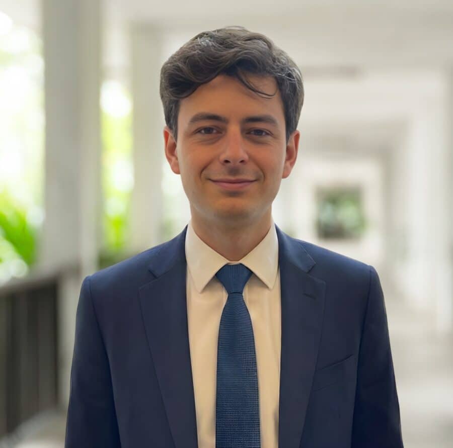 A man in a blue suit and tie stands in a bright, hallway-like setting, facing the camera and smiling slightly.