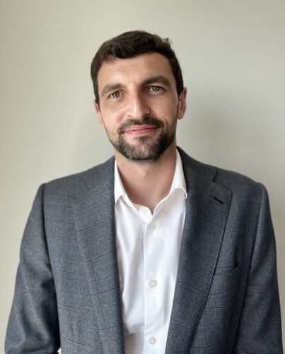 Man with short dark hair and beard wearing a grey suit jacket over a white shirt, standing in front of a plain light-coloured background.