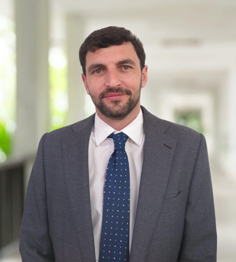 A man with short dark hair and a beard, wearing a gray suit, white shirt, and blue polka dot tie, stands in a brightly lit hallway.