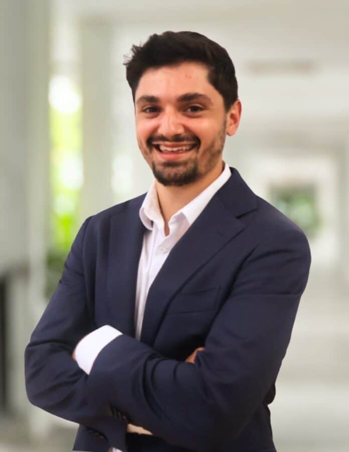 A man with short dark hair and a beard, wearing a navy suit and white shirt, stands with arms crossed, smiling in a bright, blurred indoor setting.
