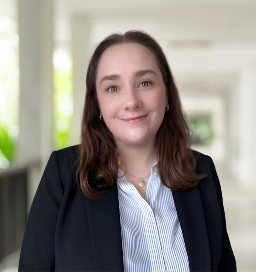 Sophia Minns, with shoulder-length brown hair, wearing a dark blazer and striped shirt, stands in a bright hallway, looking at the camera and smiling.