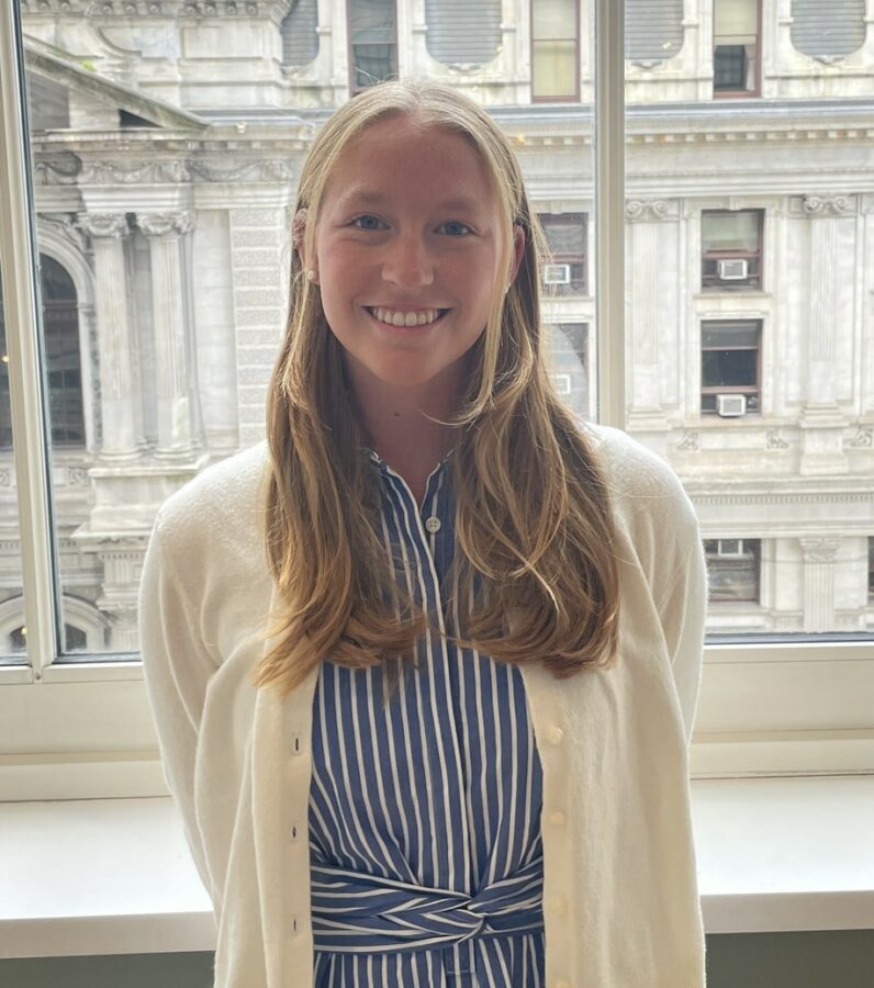 A woman with long blonde hair, wearing a blue and white striped dress and a white cardigan, stands smiling in front of a large window with a building visible outside.