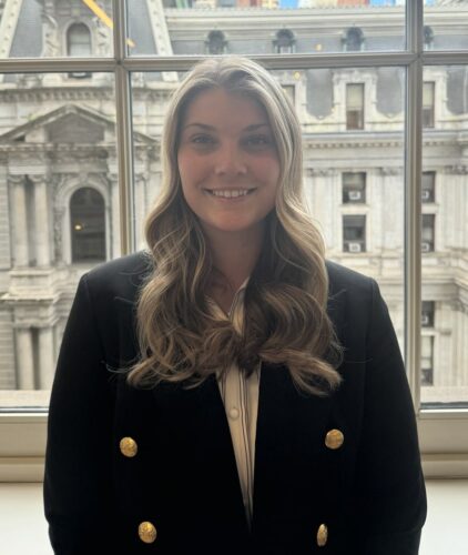 A woman with long wavy hair in a black blazer stands indoors in front of a window, with a historic stone building visible in the background.