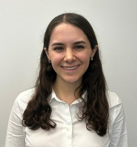 A woman with long brown hair, wearing a white button-up shirt, stands in front of a plain light-coloured wall and smiles at the camera.