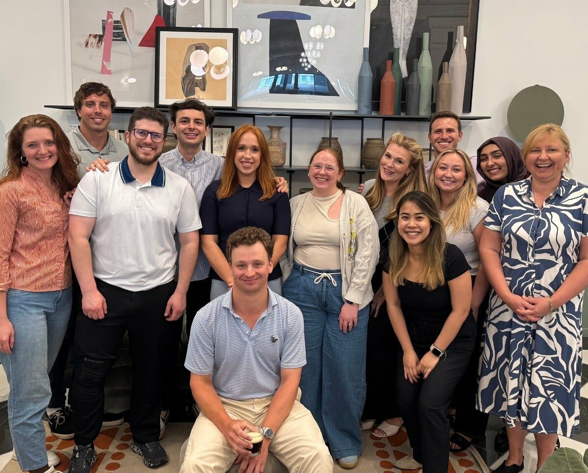 Thirteen people pose and smile together indoors in front of a decorated wall with framed art and vases, offering a glimpse of the camaraderie you’ll find when you join our dynamic team.