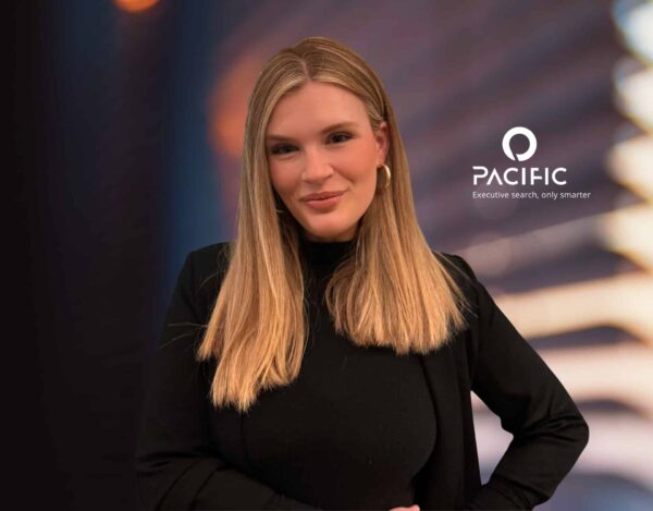 A woman with long blonde hair in a black outfit stands in front of a blurred background with the Pacific company logo and the tagline "Executive search, only smarter" on the right.