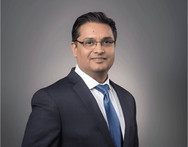 Sameer Pagnis, wearing glasses, a dark checked suit, white shirt, and blue tie, poses for a professional studio portrait against a grey background.
