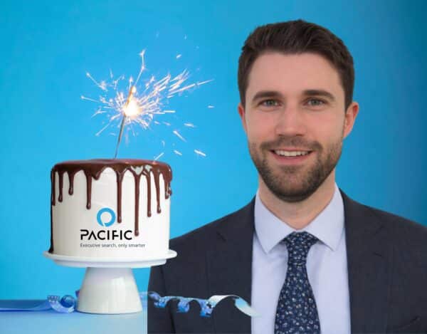 A man in a suit stands smiling next to a white cake with chocolate drizzle, a sparkler on top, and the Pacifico logo on the front—celebrating a decade of growth against a blue background.