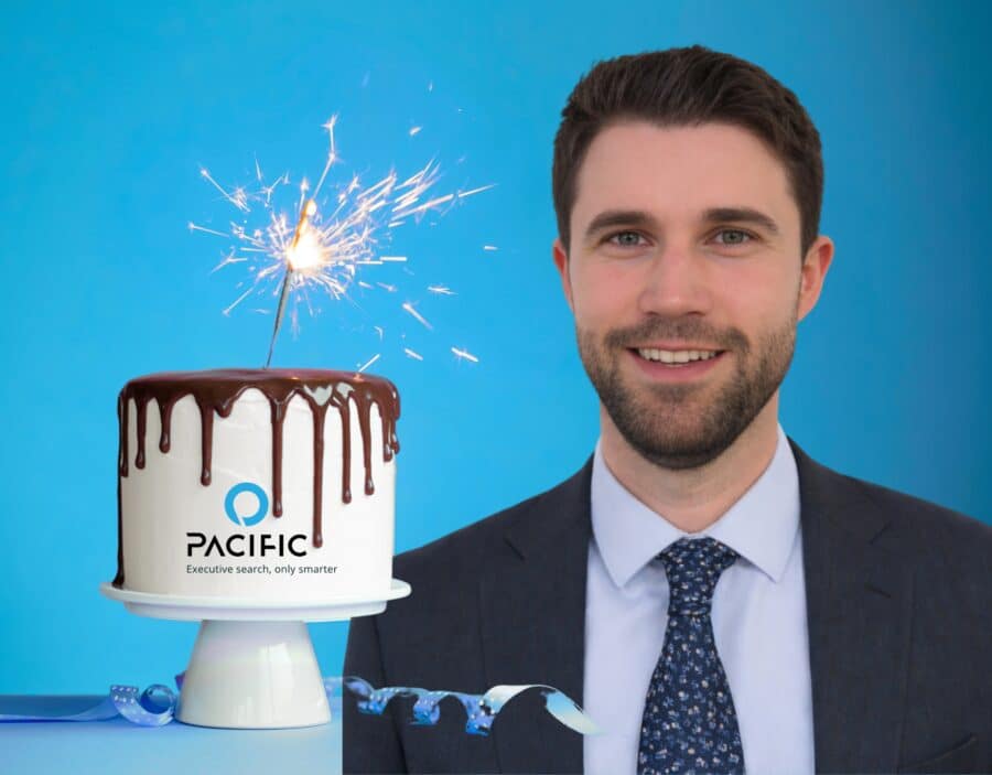 A man in a suit stands smiling next to a white cake with chocolate drizzle, a sparkler on top, and the Pacifico logo on the front—celebrating a decade of growth against a blue background.
