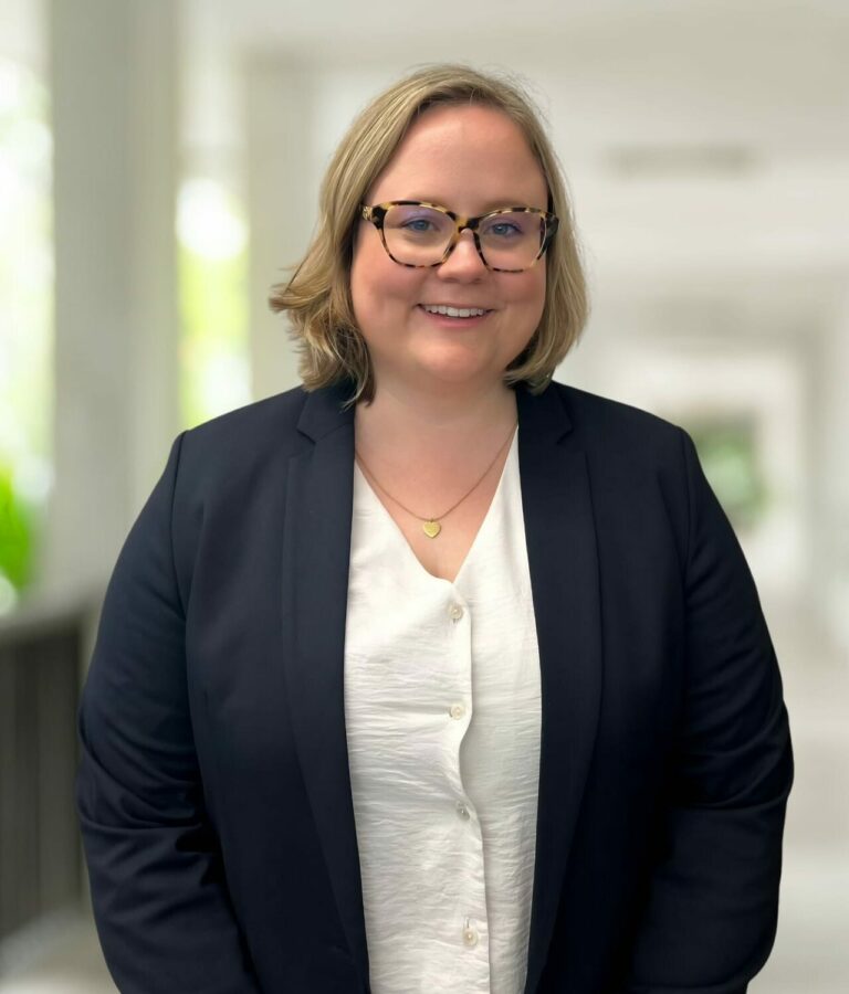 A person with shoulder-length blonde hair and glasses, wearing a dark blazer over a white blouse, stands in a bright corridor with blurred background.