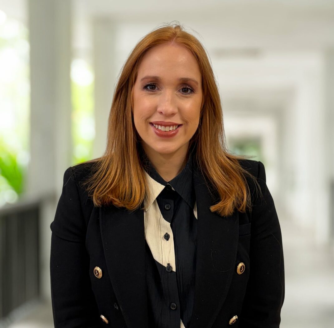 A woman with straight red hair wearing a black blazer and collared shirt stands indoors in a bright hallway, smiling at the camera—inviting you to join our dynamic team.