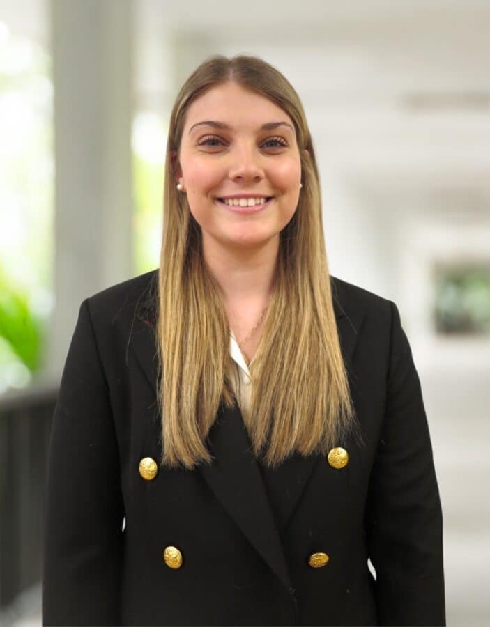 A young woman with straight blonde hair wears a black blazer with gold buttons, standing indoors with a blurred background.
