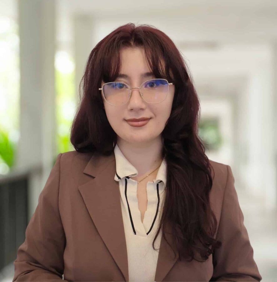 A person with long brown hair and glasses, wearing a brown blazer and a light-coloured blouse, stands in a bright indoor corridor.