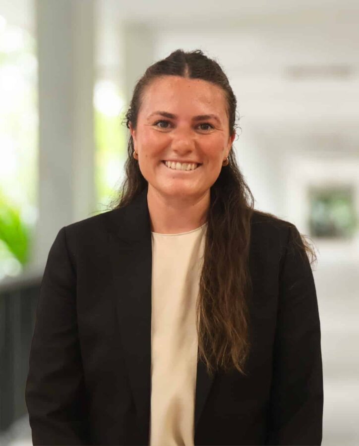 A woman with long brown hair wearing a black blazer and cream top stands indoors, smiling at the camera. The background is softly blurred.
