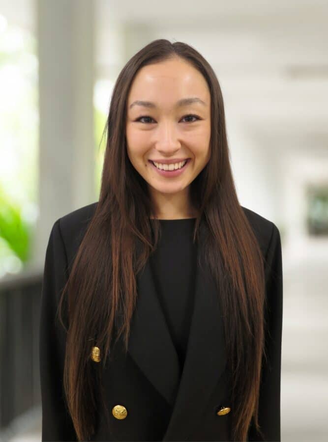 Woman with long straight brown hair wearing a black blazer stands indoors, smiling at the camera. The background is softly blurred.