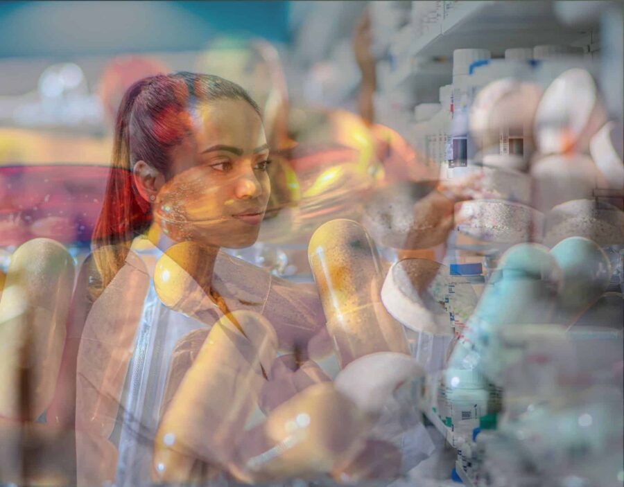 A pharmacist in a white coat reaches for a bottle on a shelf in a double-exposure image with oversized medicine capsules.